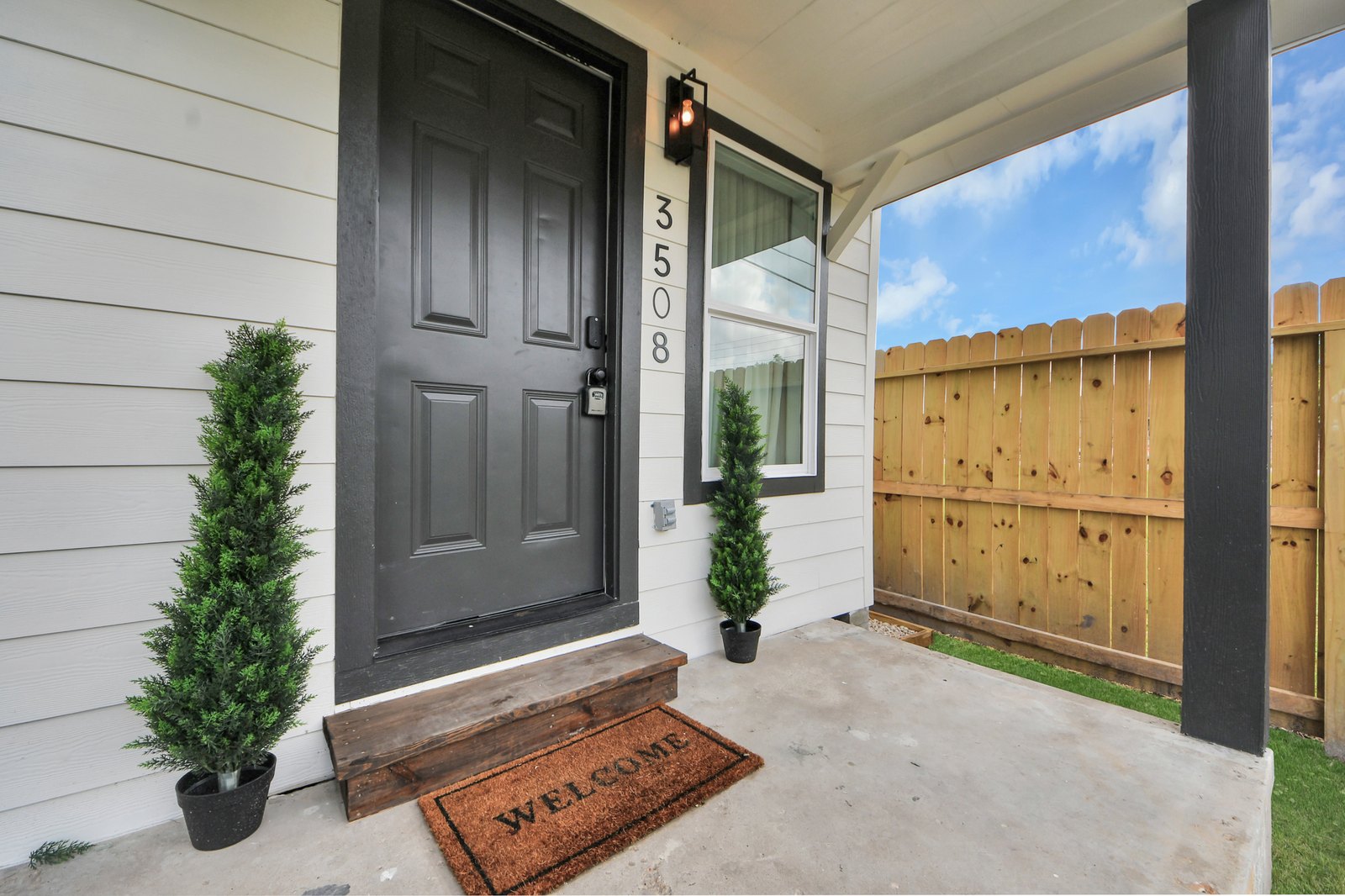 Black front door marked 3508 flanked by topiaries, with a welcome mat and keypad smart lock