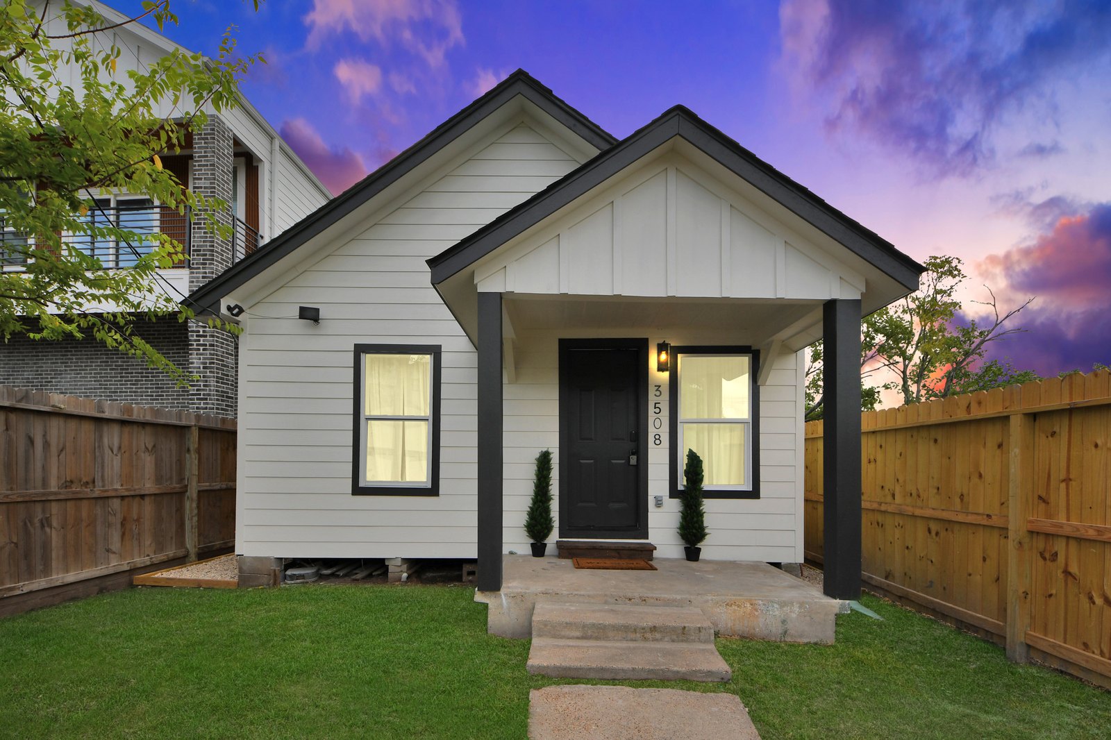 White board-and-batten bungalow with black trim, a covered front porch, and a fenced yard at 3508 Omega Street at dusk